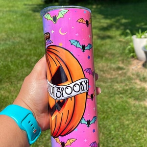 May include: A Halloween-themed tumbler with a pumpkin design and the words "Spooky". The tumbler is cylindrical, with a pink and purple gradient background, and features bats, stars, and crescent moons.