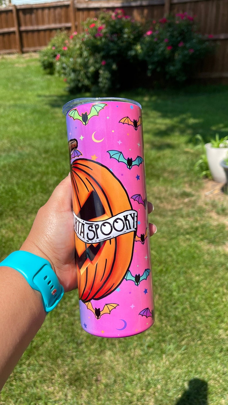 May include: A Halloween-themed tumbler with a pumpkin design and the words "Spooky". The tumbler is cylindrical, with a pink and purple gradient background, and features bats, stars, and crescent moons.