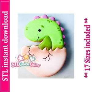 May include: A cookie shaped like a green dinosaur hatching from a light pink egg with brown crack details. The dinosaur has purple spikes along its back and a smiling face. The text STL instant download and 17 Sizes included is visible.