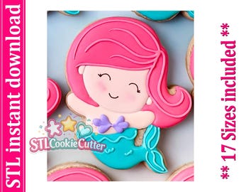 May include: A cookie shaped like a mermaid with bright pink hair, a light skin tone, and a turquoise tail. The mermaid has a purple seashell top and a smiling face. The image includes the text STL instant download and 17 Sizes included.