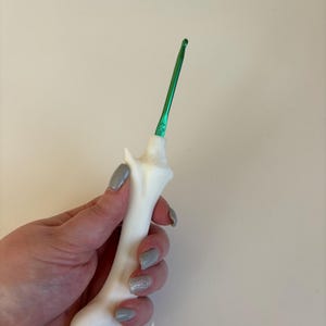 May include: A white crochet hook with a green metal hook end. The handle is a bone-shaped design. The hook is held in a hand with gray nail polish.