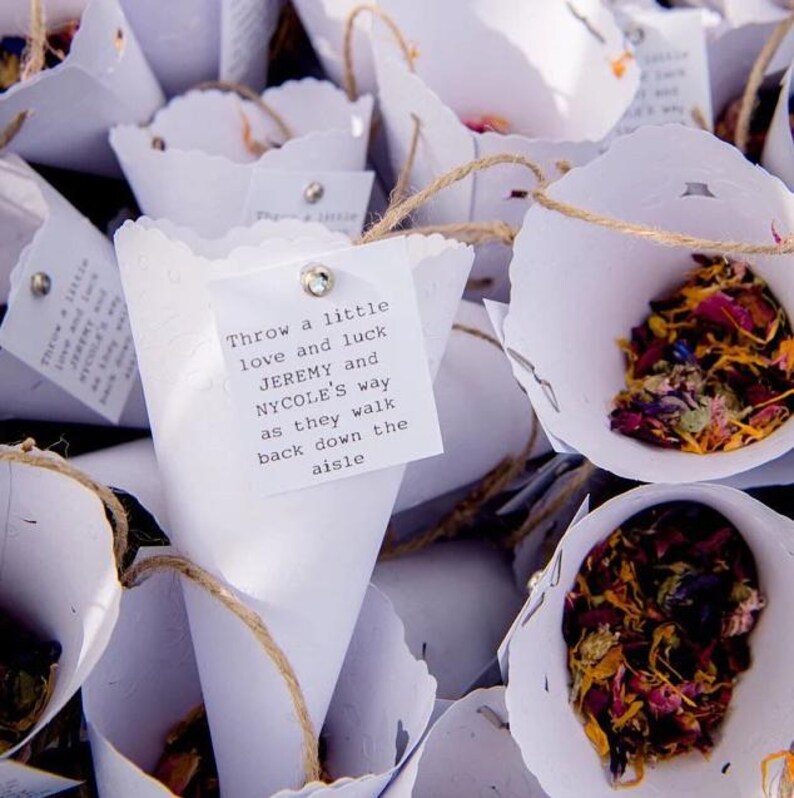 a close-up view of several white paper cones or cups filled with colorful flower petals or confetti, tied together with twine. There is a handwritten note attached to one of the cones, which reads "throw a little love and luck jEREMY and NYCOLE'S way as they walk back down the aisle".