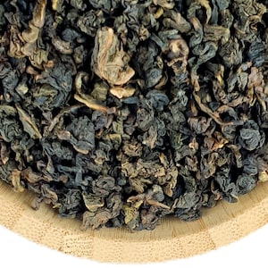 Könnte beinhalten: Eine runde, helle Holzschale gefüllt mit einem Haufen dunkler, fest gerollter und gekräuselter getrockneter Teeblätter. Die Blätter weisen eine Mischung aus tiefen Waldgrün-, Braun- und Anthrazittönen auf und wirken strukturiert.