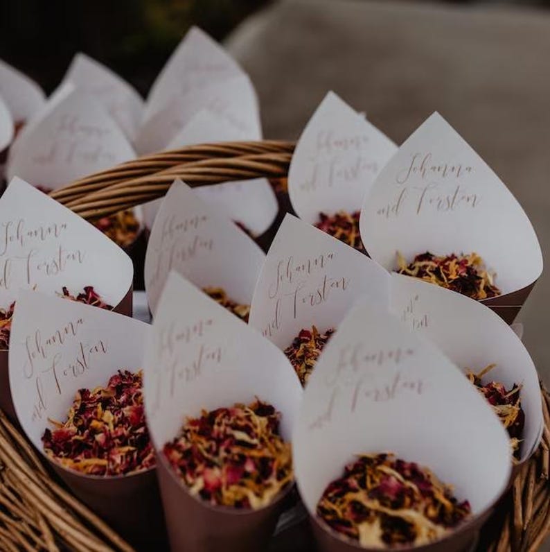 A basket filled with white paper cones containing red and yellow flower petals, with each cone labeled with the names "Schwab and Fustar".