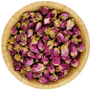 Op de afbeelding: Een houten kom gevuld met gedroogde rozenknoppen. De knoppen zijn diep roze van kleur met groene kelkbladen. De kom is gemaakt van lichtgekleurd hout en staat tegen een witte achtergrond.