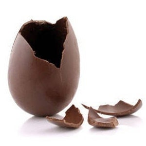 Puede incluir: Un huevo de chocolate hueco con una grieta en el lado, revelando el interior vacío. El huevo está rodeado de varios trozos de chocolate rotos.