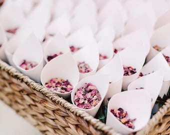 Può includere: Un cesto in vimini intrecciato riempito con numerosi coni di carta bianca, ciascuno contenente un mix di petali di fiori essiccati rosa, viola e rossi. I coni sono disposti in file all'interno del cesto, creando un'esposizione uniforme di coriandoli floreali.
