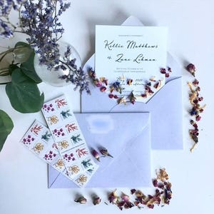 a wedding invitation with floral decorations, including a vase of purple flowers, rose petals, and a white envelope.