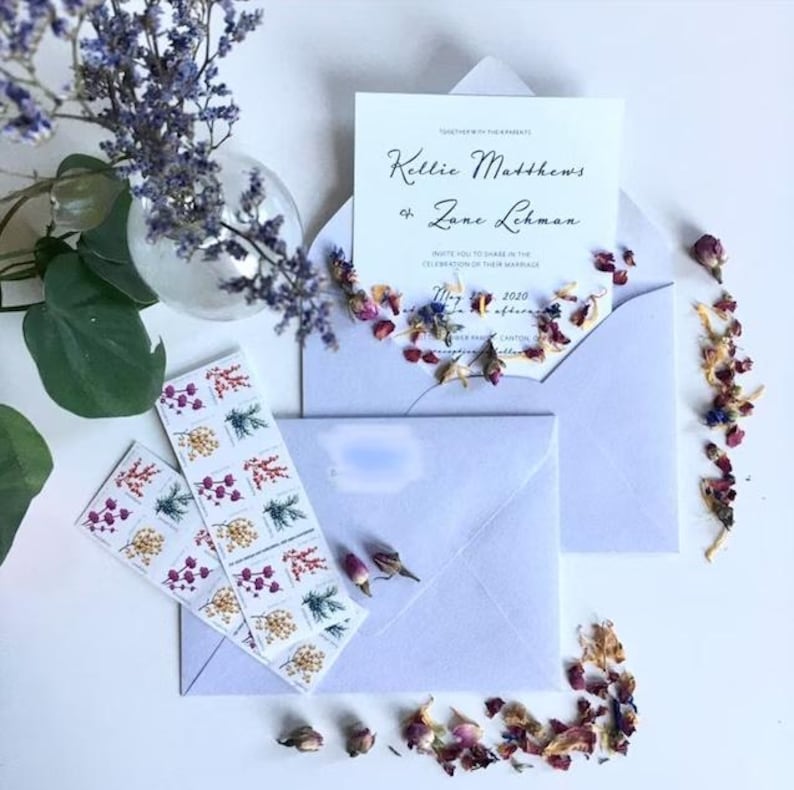 a wedding invitation with floral decorations, including a vase of purple flowers, rose petals, and a white envelope.