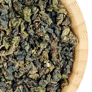 Könnte beinhalten: Eine runde, helle Bambusschale gefüllt mit einer großen Menge getrockneter, gerollter Oolong-Teeblätter. Die Teeblätter weisen eine Mischung aus dunkelgrünen, olivfarbenen und bräunlichen Tönen auf und haben ein eng gerolltes, strukturiertes Aussehen.