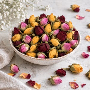 Könnte beinhalten: Eine Keramikschale gefüllt mit einer Sammlung getrockneter Rosenknospen in den Farben Dunkelrot, leuchtendem Gelb und zartem Rosa. Die Schale ist mit diesen konservierten Blütenknospen gefüllt, wobei einige lose Knospen und Blütenblätter daneben liegen.