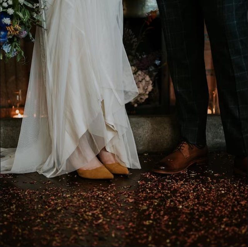 A bride and groom stand together on a wooden floor, with the bride wearing a white dress and the groom wearing brown shoes.