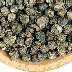 May include: A light brown wooden bowl filled with tightly rolled, dark green and silver-toned tea leaves. The leaves are shaped into small, spherical pellets that display a layered, coiled texture throughout the entire collection of dried tea.