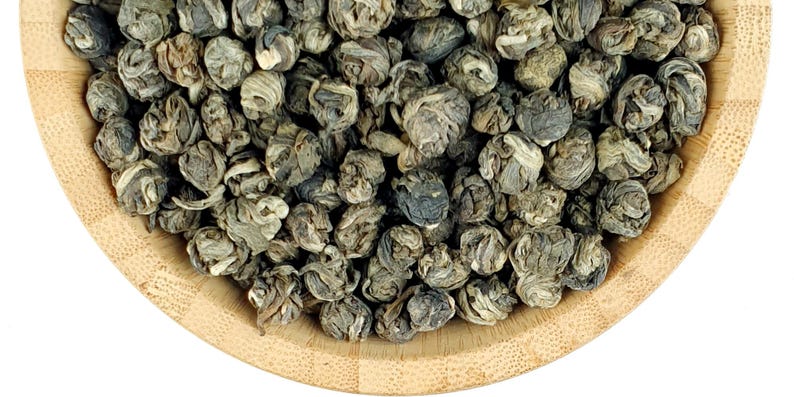 May include: A light brown wooden bowl filled with tightly rolled, dark green and silver-toned tea leaves. The leaves are shaped into small, spherical pellets that display a layered, coiled texture throughout the entire collection of dried tea.