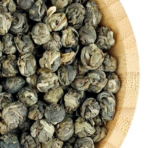 May include: A bamboo bowl filled with tightly rolled, dried green tea pearls. The tea leaves exhibit a mix of muted olive green, sage, and pale silver-white tones, showcasing a layered, spherical structure characteristic of hand-rolled tea.
