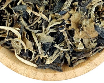 May include: A round bamboo bowl filled with loose leaf tea. The tea consists of a mixture of dark brown, nearly black, dried tea leaves and pale, light green, elongated tea buds. The texture of the leaves is crinkled and varied in size.