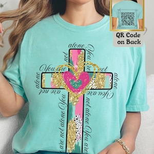 May include: A turquoise blue t-shirt with a gold glitter cross design. The cross has a pink heart in the center and the words "You are not alone You are not alone You are not alone" written around it.