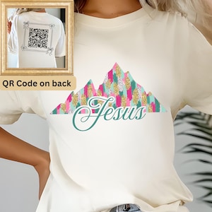 May include: A white t-shirt with a colorful mountain design and the word "Jesus" in a cursive font. The back of the shirt has a QR code.