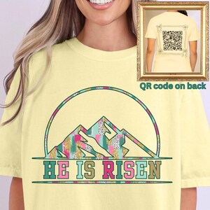 May include: A yellow t-shirt with a colorful mountain design and the text "HE IS RISEN". The back of the shirt has a QR code.