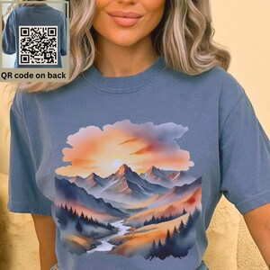 May include: A blue t-shirt with a watercolor painting of a mountain range and a sunset. The painting is in shades of orange, yellow, blue, and green. The t-shirt has a QR code on the back.
