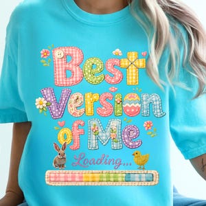 May include: A light blue t-shirt with the text "Best Version of Me" in colorful patchwork letters. The design includes a cross, an Easter egg, flowers, a bunny, a chick, and a loading bar.