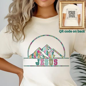 May include: A white t-shirt with a green, pink, and gold mountain design and the word "JESUS" printed below. The back of the shirt features a QR code.