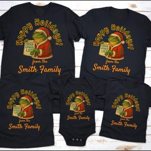 May include: A collection of black t-shirts and a onesie featuring a frog dressed as Santa Claus. The frog holds a "Croak-Approved List" and the text reads "Happy Holidays!" and "from the Smith Family" in gold lettering.