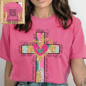 May include: A pink t-shirt with a cross design featuring a heart and the words "You are not alone." The cross has gold glitter and colorful accents. A smaller image shows the back of the shirt with a QR code.
