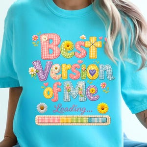May include: A light blue t-shirt with the text "Best Version of Me" in colorful, patchwork-style letters. The design includes flowers, hearts, and a loading bar graphic, creating a cheerful and positive vibe.