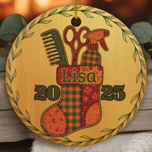 May include: A round, yellow ceramic Christmas ornament featuring a stocking design with a comb, scissors, and spray bottle. The name "Lisa" is displayed above the numbers "2025". The ornament is trimmed with a green leaf design and a gold string for hanging.