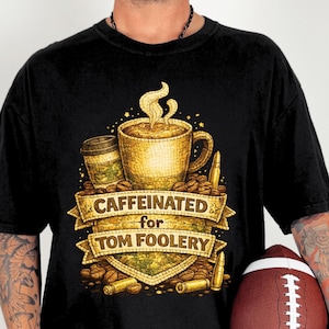 May include: Black t-shirt with a coffee-themed graphic. The design features a golden coffee cup, a to-go cup, coffee beans, and the text "CAFFEINATED for TOM FOOLERY" on a banner. Two bullets are on either side of the banner.