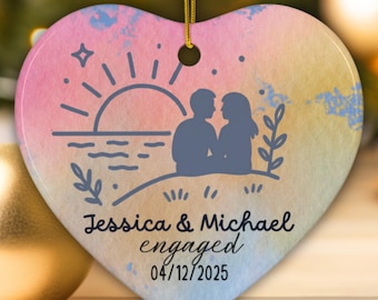 May include: Heart-shaped ceramic ornament with a watercolor sunset design. Features a silhouette of a couple, the text "Jessica & Michael engaged 04/12/2025", and the word "PERSONALIZABLE". Includes a gold string for hanging.