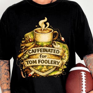 May include: Black t-shirt with a graphic featuring a coffee cup, fishing lures, and a fish. The text on the shirt reads "Caffeinated for Tom Foolery". A football is held in the person's hand.