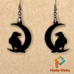May include: A pair of black acrylic earrings featuring a crescent moon with a raven silhouette on each earring. The earrings have silver hooks.