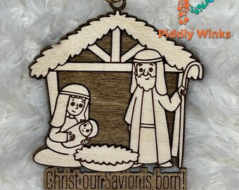 May include: Wooden Christmas ornament depicting the Nativity scene. The ornament features Joseph, Mary, and baby Jesus in a manger, with the text "Christ our Savior is born!" The ornament has a light wood color with darker wood accents.