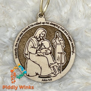 May include: A wooden ornament with a depiction of Jesus healing a man. The ornament is brown and features a white background with the text "Instantly, the man was healed! He rolled up his sleeping mat and began walking..." and "John 5:9".