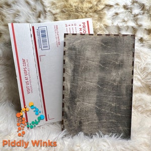 May include: A wooden box with a dark gray stain finish. The box has a slightly distressed look and is made of wood with a visible grain pattern. The box is sitting on a white fluffy surface.