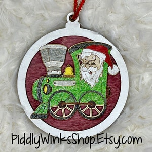May include: A round wooden Christmas ornament with a white border. The ornament features a green train with a red-hatted Santa Claus riding in the front. The train has a bell and a nameplate. The background is a red and white woodgrain pattern.