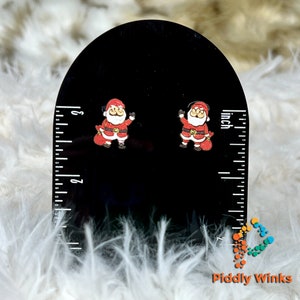 May include: Santa Claus stud earrings. These festive earrings feature a red and white Santa Claus design with a black background. The earrings are displayed on a black ruler, with a measurement of approximately 1 inch.
