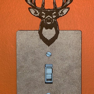 Puede incluir: Una tapa de interruptor de luz con un acabado de madera marrón y una silueta de cabeza de ciervo en la parte superior. El interruptor es un interruptor de palanca estándar con un botón blanco.