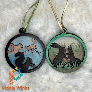 May include: Two round wooden ornaments with a nature theme. One ornament features a blue and brown background with a silhouette of two squirrels. The other ornament features a green and brown background with a silhouette of two rabbits.