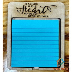 May include: Wooden notepad holder with a blue lined notepad. The holder has the text "It takes a big heart to teach little minds." with two hearts on either side of the word "heart".