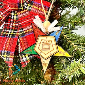 Puede incluir: Adorno navideño de madera en forma de estrella, con segmentos coloridos y un emblema central. El adorno cuelga de un árbol de Navidad, con un lazo escocés rojo en el fondo.