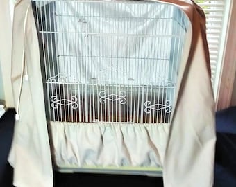 May include: A white metal birdcage with a decorative design on the front, draped with a beige fabric cover. The cover is tied at the top and has a ruffled skirt at the bottom, creating a cozy and elegant look.