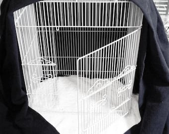 May include: A white wire bird cage with an open door, partially covered by a dark blue fabric. The cage sits on a white surface, with a decorative design on the bottom.