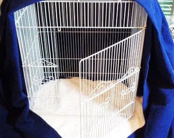 May include: A white wire birdcage with an open door is partially covered by a dark blue fabric cover. The cage sits on a white, textured surface. The cage has a decorative design on the bottom.