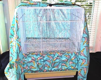 May include: A white metal birdcage covered with a turquoise fabric featuring a bird pattern. The fabric drapes over the cage, creating a cozy environment. The cage sits on a light yellow base, with a window in the background.