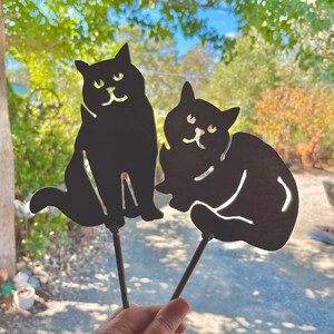 May include: Two black metal cat silhouettes on sticks. The cats are facing forward with their tails curled around their legs. The cats are about 25 cm tall.