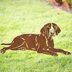 May include: A rusty brown metal silhouette of a German Shorthaired Pointer dog lying down. The dog is facing the camera with its head turned slightly to the side. The silhouette is placed on a green grassy lawn.