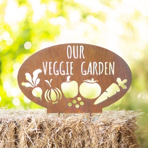 May include: A rusty metal sign with the words "Our Veggie Garden" in white lettering. The sign is shaped like a garden with cutouts of vegetables, including a carrot, tomato, onion, and radish.